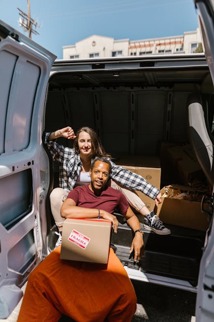 Two women are loading cardboard boxes into the rear compartment of a panel van, associated with house removals in Croydon. The woman in the foreground, dressed in a maroon shirt and sitting on a red b