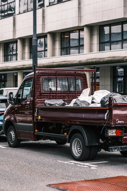 Two women are loading cardboard boxes into the rear compartment of a panel van, associated with house removals in Croydon. The woman in the foreground, dressed in a maroon shirt and sitting on a red b