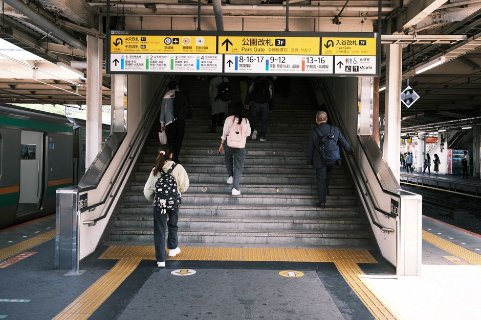 A group of people, including adults and a child, are walking up a staircase inside a train station, preparing for a home relocation or transfer to another part of the station. The staircase, made of g