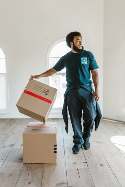 A professional mover from Removals Croydon, dressed in a dark blue t-shirt and trousers, is inside a well-lit, empty room with light-colored walls and wooden flooring, carrying a cardboard box with re