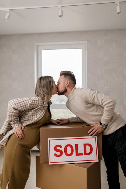 A professional mover from Removals Croydon, dressed in a dark blue t-shirt and trousers, is inside a well-lit, empty room with light-colored walls and wooden flooring, carrying a cardboard box with re